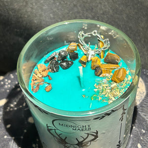 Close up image of Glass votive candle with a green candle dressed with black tourmaline and tigers eye crystal chips, oak and mugwort and a deer head charm