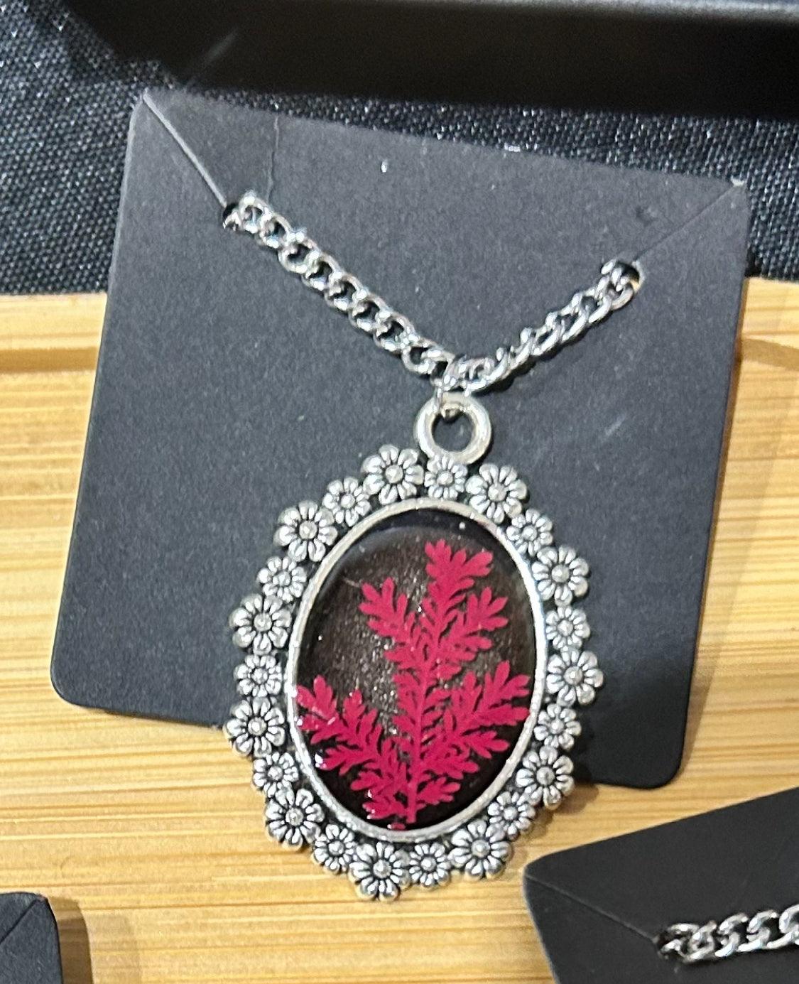 Red fern leaf necklace set on a black background and encased in a silver frame with carved flowers around it.